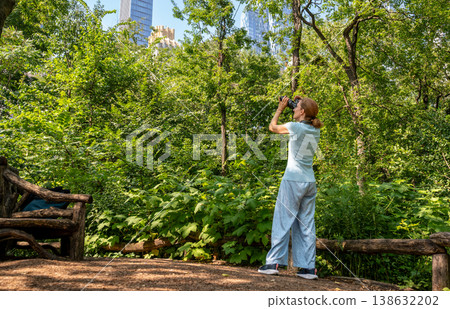 New york, usa august 14,2024. Woman enjoying outdoor leisure activity, birdwatching with binoculars in central park, surrounded by green trees and city skyline New york, usa august 14,2024. Woman enjoying outdoor leisure activity, birdwatching with binoculars in central park, surrounded by green trees and city skyline 138632202