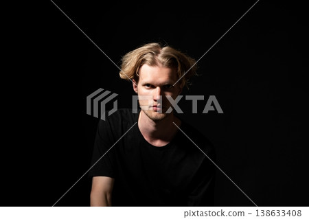 Young man in a black t-shirt facing the camera on a dark background with dramatic lighting highlighting his serious expression, conveying mystery and mood 138633408