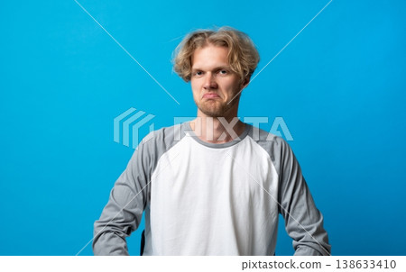 Young blond man with beard expressing skepticism and doubt with a displeased grimace, feeling unsure or disapproving against a plain blue background, showing distrust 138633410