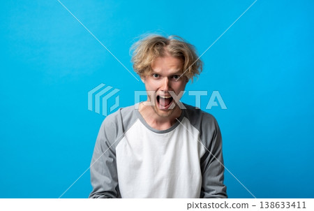 Young man screaming with wide-open mouth and messy hair, headshot conveying intense anger, frustration and emotional outburst against a blue studio background 138633411