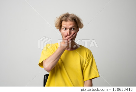 Young man covering mouth with hand, looking sideways with wide eyes, expressing shock, surprise, gossip, secret, silence, emotion, and an unexpected reaction 138633413