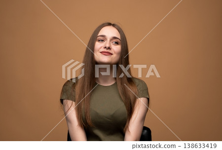 Young woman with long brown hair posing against a beige background, smiling and looking upward to the side 138633419