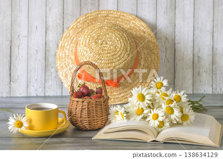 Summer still life; yellow cup of tea, bouquet of white daisy flowers on a open book, wicker basket with ripe strawberries and straw hat on a wooden background Summer still life; yellow cup of tea, bouquet of white daisy flowers on a open book, wicker basket with ripe strawberries and straw hat on a wooden background 138634129