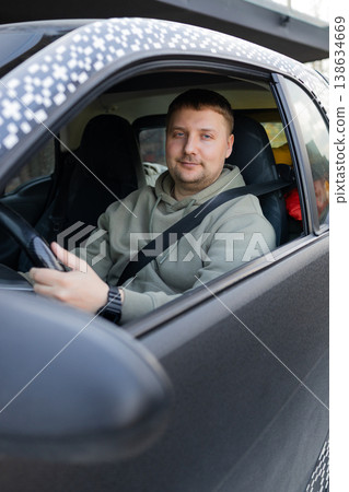 Adult man sitting behind steering wheel driving car and looking at camera, everyday transportation and lifestyle concept. Adult man sitting behind steering wheel driving car and looking at camera, everyday transportation and lifestyle concept. 138634669