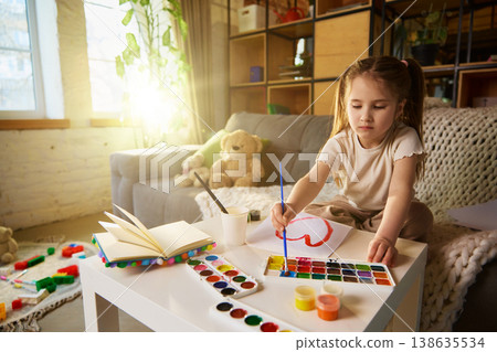 Little girl painting colorful heart with watercolor at table in cozy living room. 138635534