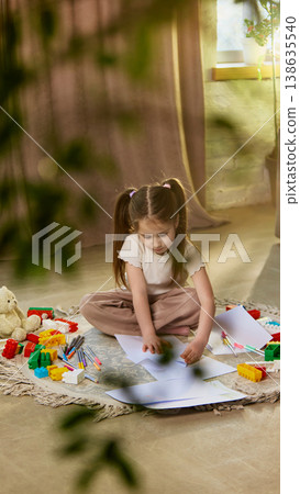 Girl sitting on floor surrounded by toys and drawings in cozy home space Girl sitting on floor surrounded by toys and drawings in cozy home space 138635540