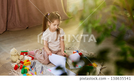 Little girl sorting drawings and papers sitting on floor with toys. 138635541