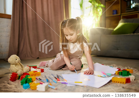 Little girl drawing on paper on floor with colorful toys in warm sunlight. 138635549