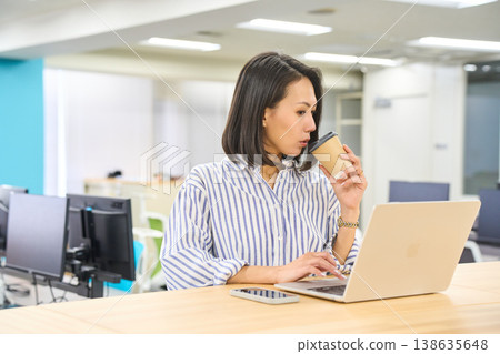 一位女士在辦公室的站立式辦公桌前使用筆記型電腦工作,手裡拿著一杯咖啡。 一位女士在辦公室的站立式辦公桌前使用筆記型電腦工作,手裡拿著一杯咖啡。 138635648