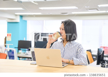 A woman working on her laptop at a standing desk in the office, holding a cup of coffee. A woman working on her laptop at a standing desk in the office, holding a cup of coffee. 138635651