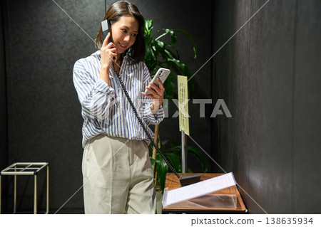 A woman at the office reception desk makes an internal phone call while checking her smartphone. 138635934
