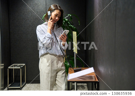 A woman at the office reception desk makes an internal phone call while checking her smartphone. 138635936