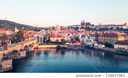 The sun rises gently over Prague, illuminating the Vltava River and creating a warm glow on the rooftops. 138637961