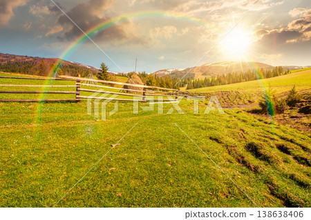 sunset over mountainous countryside landscape in spring. wooden fence and grassy meadow on a green hill. distant mountain ridge with snow capped tops. cloudy sky and rainbow sunset over mountainous countryside landscape in spring. wooden fence and grassy meadow on a green hill. distant mountain ridge with snow capped tops. cloudy sky and rainbow 138638406