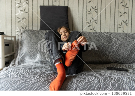 A young girl sitting on a patterned sofa and putting on bright orange socks, preparing to go out. 138639551