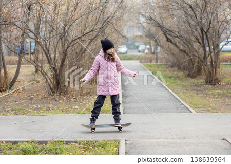 Full length portrait of a young girl standing on a skateboard in the middle of an autumn park alley. Full length portrait of a young girl standing on a skateboard in the middle of an autumn park alley. 138639564
