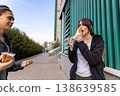 Close up of a young girl with a backpack eating a sandwich from a plastic box while talking to a friend. 138639585