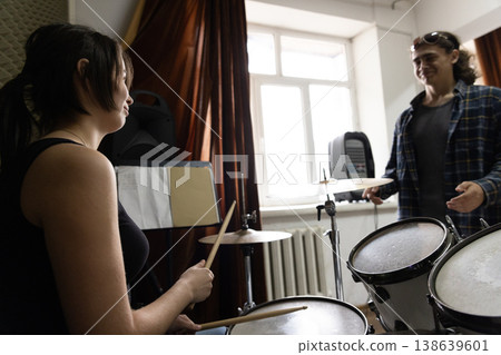 A young female music student with drumsticks looking at her male colleague during a practice session in a studio. A young female music student with drumsticks looking at her male colleague during a practice session in a studio. 138639601