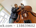 Dynamic low angle view of a young college student playing a large wooden double bass in a bright music room. 138639602