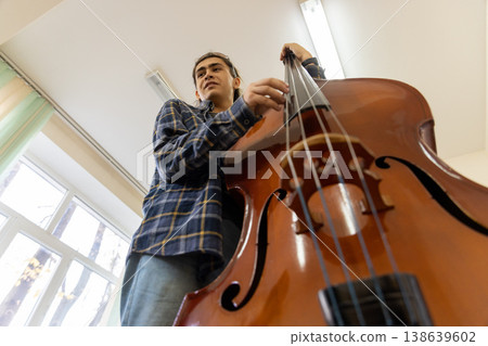 Dynamic low angle view of a young college student playing a large wooden double bass in a bright music room. 138639602