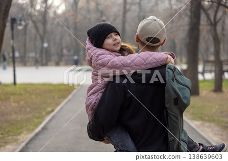 A father walks through a park carrying his daughter in a pink coat, who is hugging him from behind and smiling. 138639603