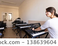 Young woman adjusting a synthesizer in a study hall filled with various musical keyboards. 138639604