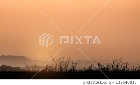 A serene landscape featuring the silhouette of grass in the foreground and misty hills under a warm orange morning sky A serene landscape featuring the silhouette of grass in the foreground and misty hills under a warm orange morning sky 138640833