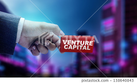 Professional hand holding a red block with Venture Capital text. Background is a blurred urban night scene with neon lights, representing corporate growth and startup funding 138640843