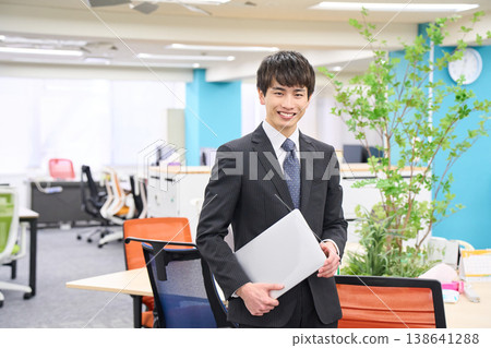 A young businessman with a laptop in the office, sporting a cheerful smile. A young businessman with a laptop in the office, sporting a cheerful smile. 138641288