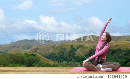 A young Indonesian southeast asian muslim woman in a hijab and modest sportswear practices yoga and stretching on a red mat with a stunning mountain landscape background A young Indonesian southeast asian muslim woman in a hijab and modest sportswear practices yoga and stretching on a red mat with a stunning mountain landscape background 138641329