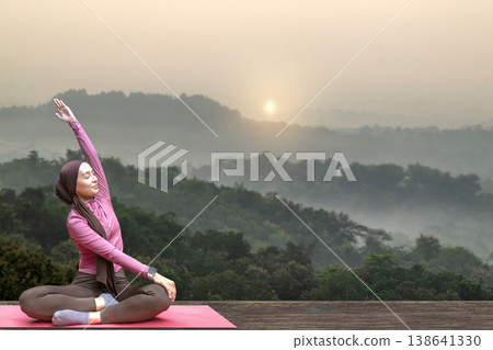 A young Indonesian southeast asian muslim woman in a hijab practices yoga and stretching on a mat outdoors against a beautiful misty mountain landscape during sunrise. A young Indonesian southeast asian muslim woman in a hijab practices yoga and stretching on a mat outdoors against a beautiful misty mountain landscape during sunrise. 138641330