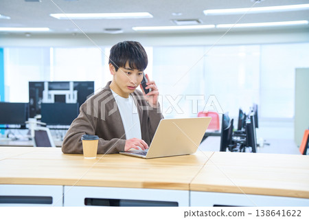 A male employee opens his laptop at a free-address desk and makes a call on his smartphone. A male employee opens his laptop at a free-address desk and makes a call on his smartphone. 138641622