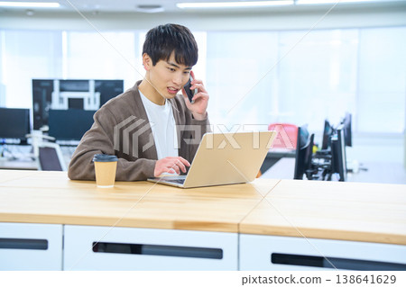A male employee opens his laptop at a free-address desk and makes a call on his smartphone. 138641629