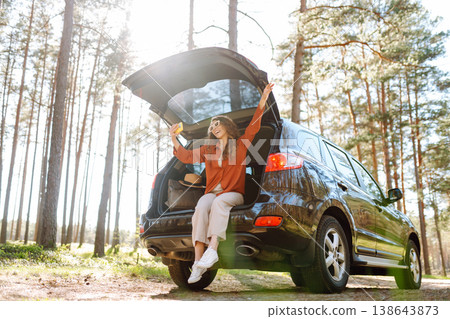 A cheerful woman sits on the trunk of her car and taking a selfie. Selfie while traveling by car. 138643873