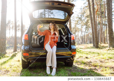 A cheerful woman sits on the trunk of her car and taking a selfie. Selfie while traveling by car. 138643887