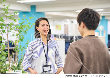 A female employee is seen smiling and talking with a male colleague in the office while holding a device. 138644575