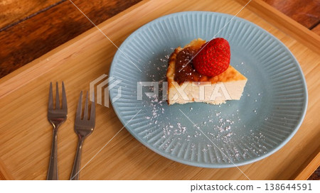 close up of a slice of basque cheesecake with a strawberry on top placed on a blue plate with white sugar powder on a wooden tray with two small forks close up of a slice of basque cheesecake with a strawberry on top placed on a blue plate with white sugar powder on a wooden tray with two small forks 138644591
