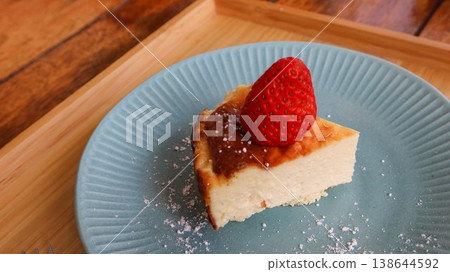 close up of a slice of basque cheesecake with a strawberry on top placed on a blue plate with white sugar powder on a wooden tray with two small forks 138644592
