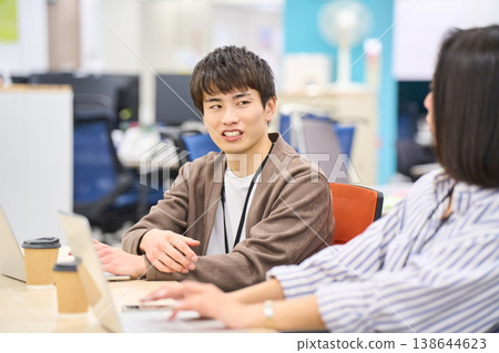 一位年輕的男員工面帶微笑，在辦公桌前與一位女同事聊天。 138644623