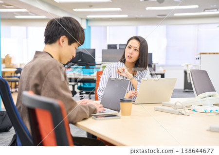 A female senior colleague smiles as she listens to a junior colleague talking about working on their computer in the office. A female senior colleague smiles as she listens to a junior colleague talking about working on their computer in the office. 138644677