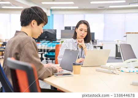 一位資深女同事面帶微笑地聽著一位年輕同事談論在辦公室用電腦工作的情況。 138644678