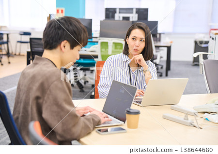一位資深女同事面帶微笑地聽著一位年輕同事談論在辦公室用電腦工作的情況。 一位資深女同事面帶微笑地聽著一位年輕同事談論在辦公室用電腦工作的情況。 138644680