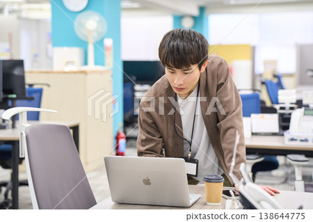 A young male employee checks his laptop screen while standing in a free-address office. A young male employee checks his laptop screen while standing in a free-address office. 138644775