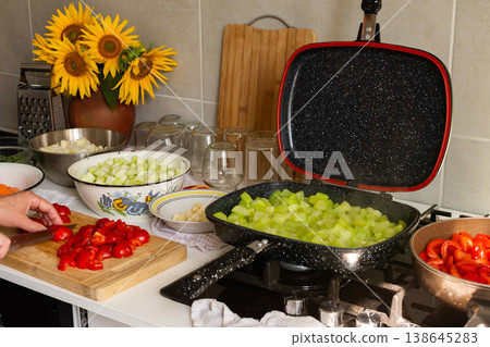A woman cuts tomatoes on a wooden board surrounded by fresh vegetables while preparing squash caviar for winter canning in her kitchen 138645283