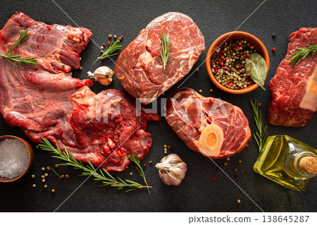 Several types of beef steak are laid out on a dark surface with fresh herbs and spices. Ingredients include garlic, pepper, and oil, showing preparation for cooking in a kitchen 138645287