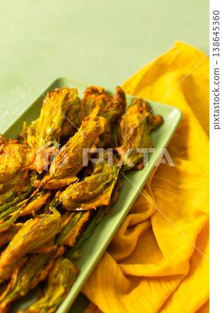 Delicious deep-fried zucchini flowers are neatly arranged on a green platter, showcasing their crispy texture. A bright yellow napkin adds warmth to the scene 138645360
