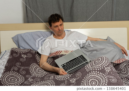 A man is sitting on a bed in a bedroom. He is wrapped in blankets and holding a laptop. He looks frustrated and tired in the early morning light A man is sitting on a bed in a bedroom. He is wrapped in blankets and holding a laptop. He looks frustrated and tired in the early morning light 138645401
