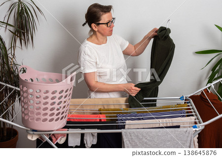 A 50-year-old woman with glasses hangs washed clothes on a drying rack in her indoor space. She sorts through the laundry from a pink basket as she prepares it to dry A 50-year-old woman with glasses hangs washed clothes on a drying rack in her indoor space. She sorts through the laundry from a pink basket as she prepares it to dry 138645876