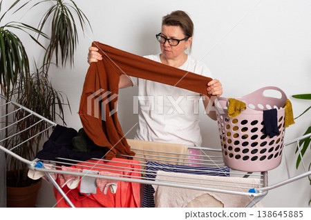 A 50-year-old woman is in her room. She is hanging washed laundry on a drying rack. A basket of clothes is nearby. She looks focused on her task A 50-year-old woman is in her room. She is hanging washed laundry on a drying rack. A basket of clothes is nearby. She looks focused on her task 138645885