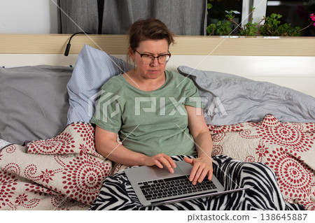 A woman sits on bed in bedroom and works on laptop. She has blankets around her and seems focused on task while enjoying her own space A woman sits on bed in bedroom and works on laptop. She has blankets around her and seems focused on task while enjoying her own space 138645887